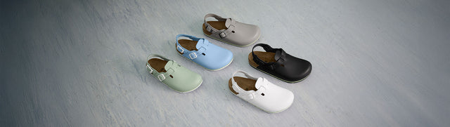 Cinque paia sabot Birkenstock Professional, ideali per uso professionale in settori come HORECA, sanitario e industriale, disposti su una superficie grigia che ricorda un pavimento. I sabot sono visti dall'alto e leggermente di lato, mettendo in risalto le diverse colorazioni e il design classico del modello.