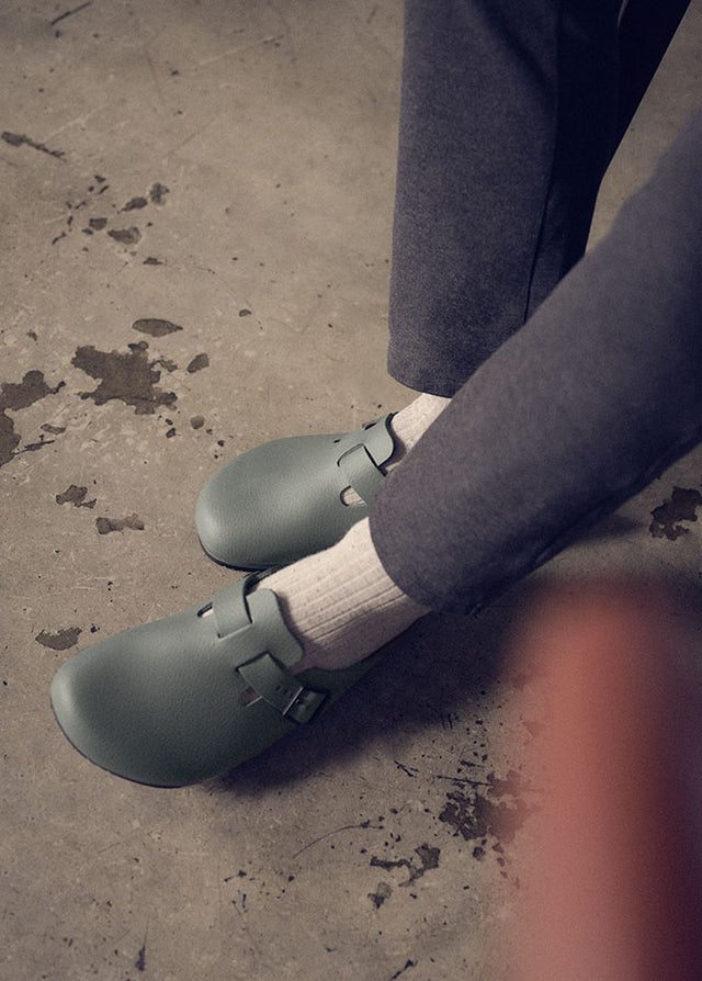 Birkenstock Boston Pro di colore thyme indossate con calzini bianchi e pantaloni scuri. Le calzature, con la loro fibbia regolabile, sono adatte per l'uso professionale in ambienti come ristoranti, hotel e altre strutture ricettive, offrendo comfort e supporto per lunghe ore in piedi.  La foto è scattata da una prospettiva leggermente dall'alto su una superficie in cemento con alcune macchie, che suggerisce un ambiente di lavoro pratico.