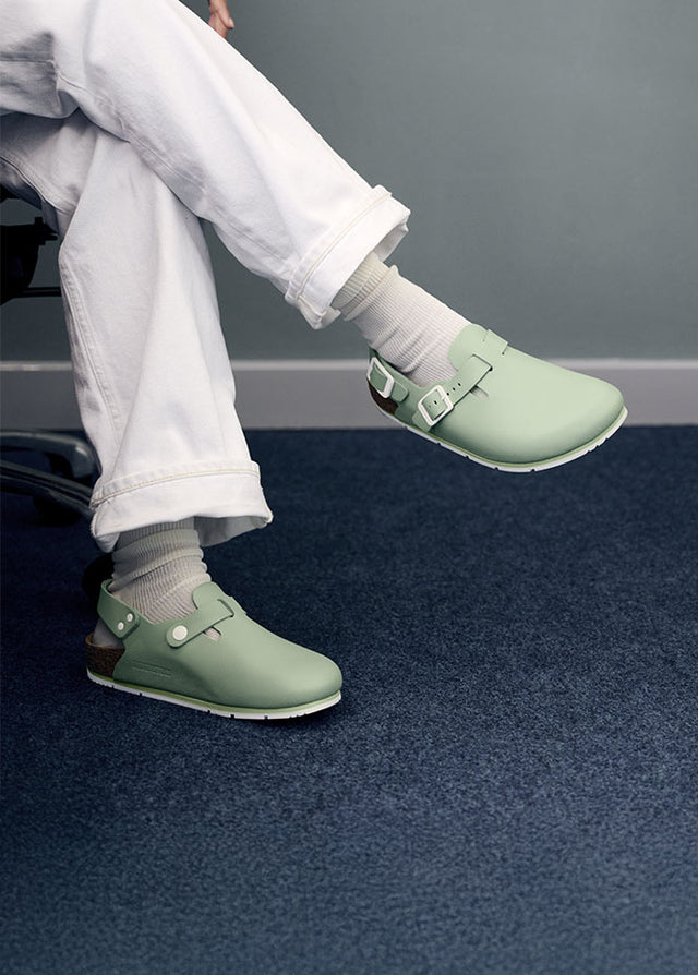 Birkenstock Professional Tokio Pro colore verde menta, con fibbie regolabili e cinturino posteriore per una maggiore stabilità. Le scarpe sono indossate con calzini bianchi e pantaloni bianchi arrotolati, su una superficie di moquette blu. La persona seduta suggerisce l'uso di queste calzature in ambienti professionali come il settore sanitario, dove il comfort, l'igiene e il supporto sono fondamentali per lunghe ore di lavoro.