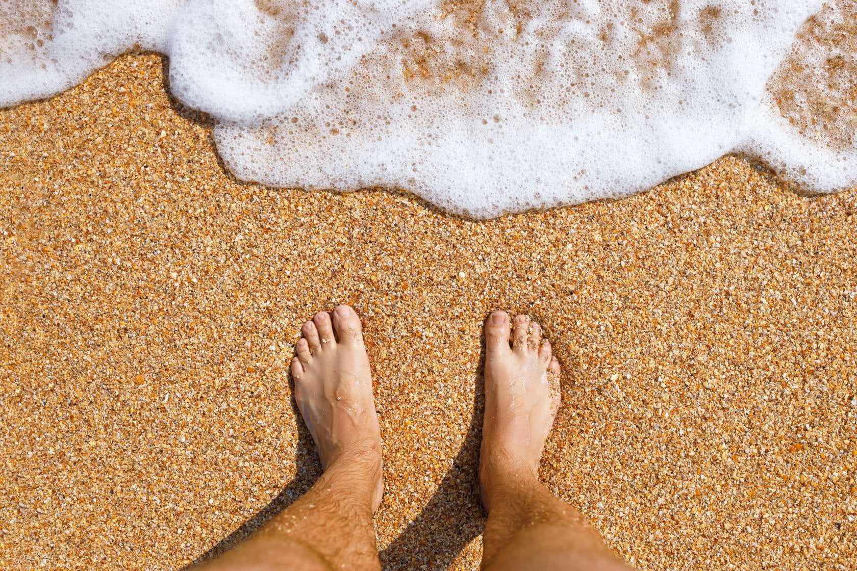 Dettaglio di piedi nudi su spiaggia granulosa, mentre le onde si infrangono sulla riva.