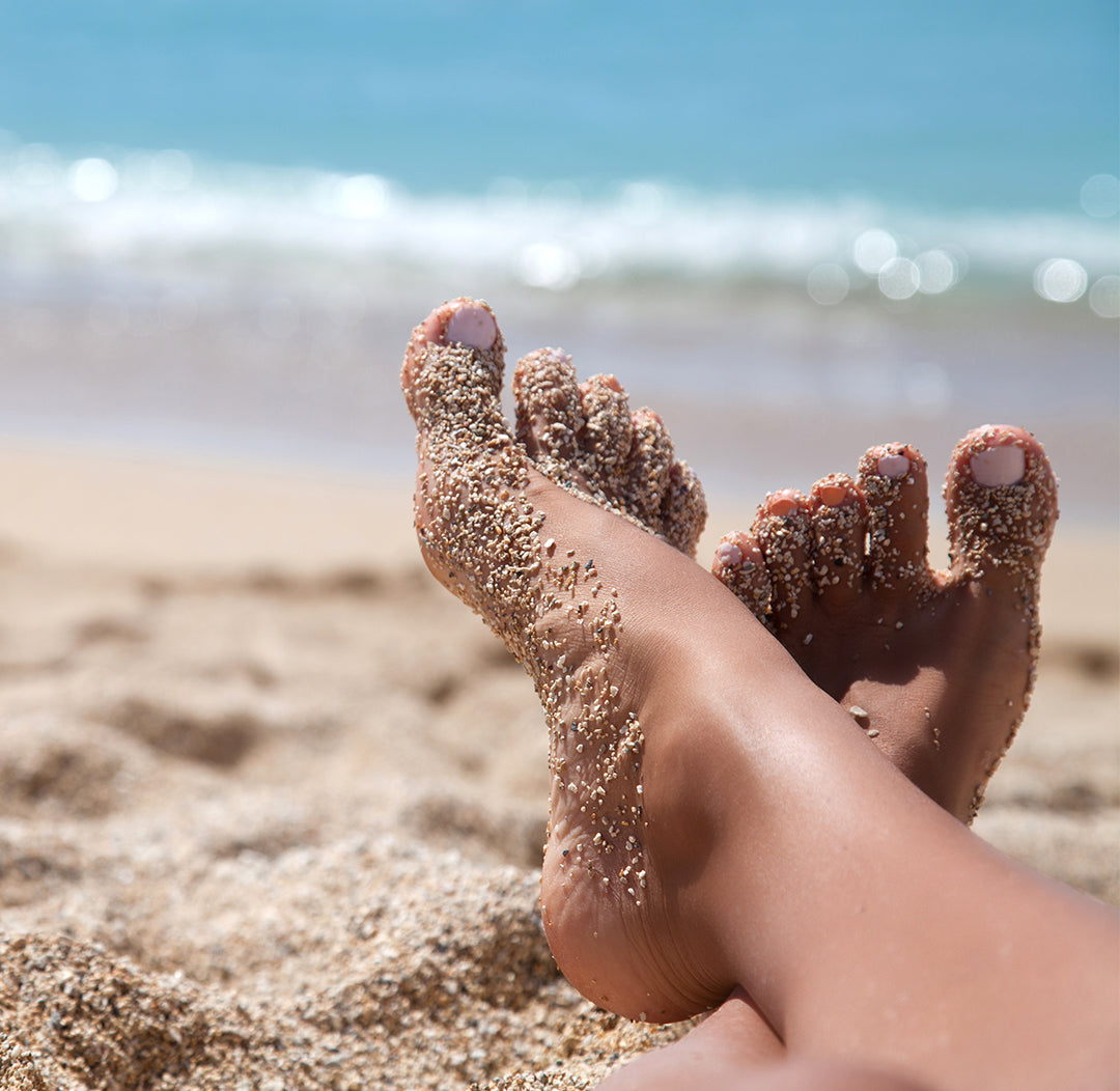 Primo piano di piedi femminili parzialmente coperti di sabbia, mare sfocato sullo sfondo.