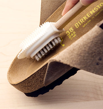 Spazzola con setole di nylon e gommini sul lato opposto che pulisce il plantare in pelle scamosciata di un sandalo Birkenstock marrone.