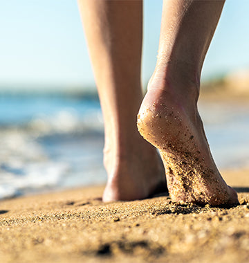 Primo piano di piedi nudi che camminano sulla sabbia, con il mare sullo sfondo.