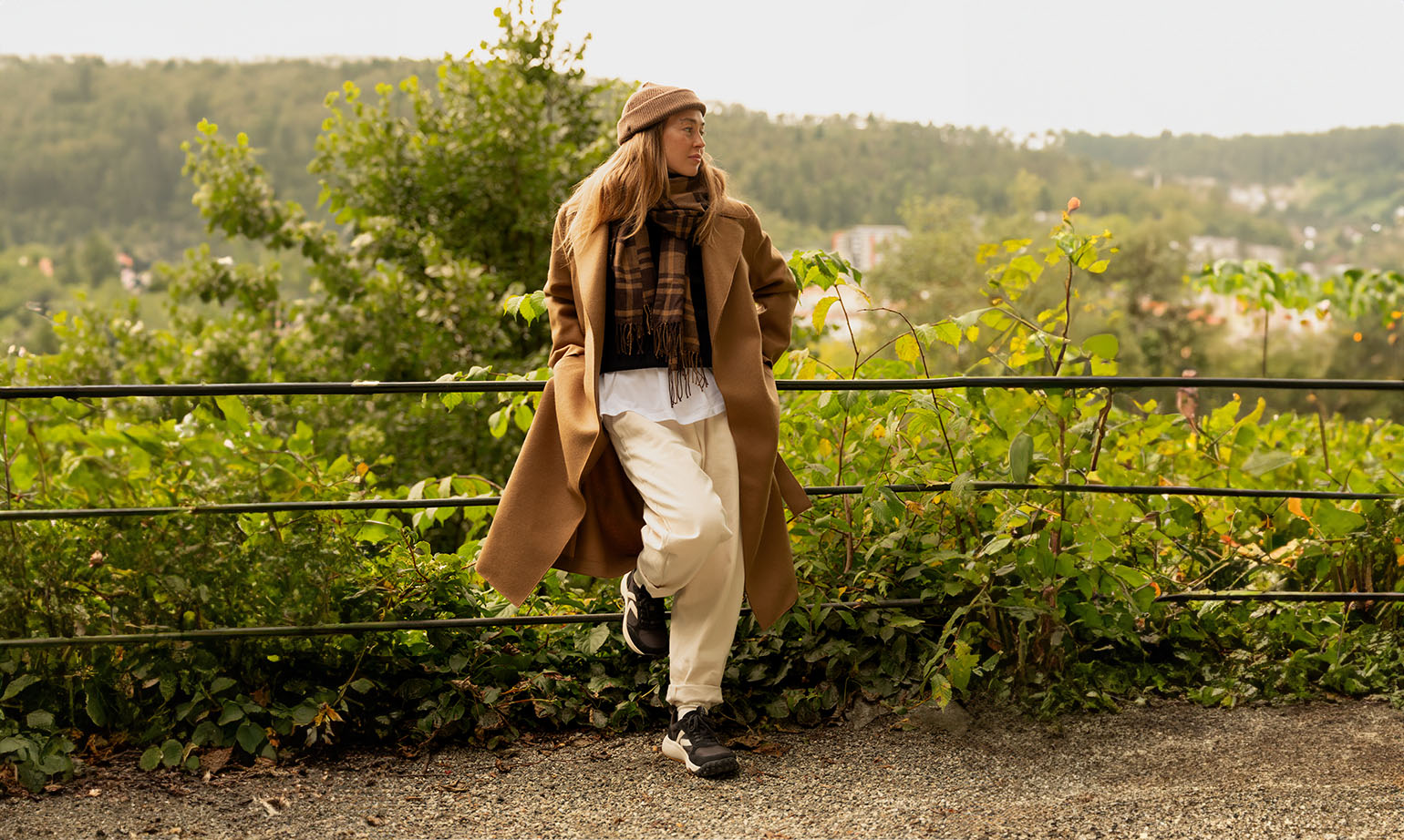 Donna che indossa sneakers KEEN KS 86 nere, cappotto cammello, pantaloni chiari e sciarpa scozzese, appoggiata a una ringhiera in un ambiente naturale con vista panoramica.