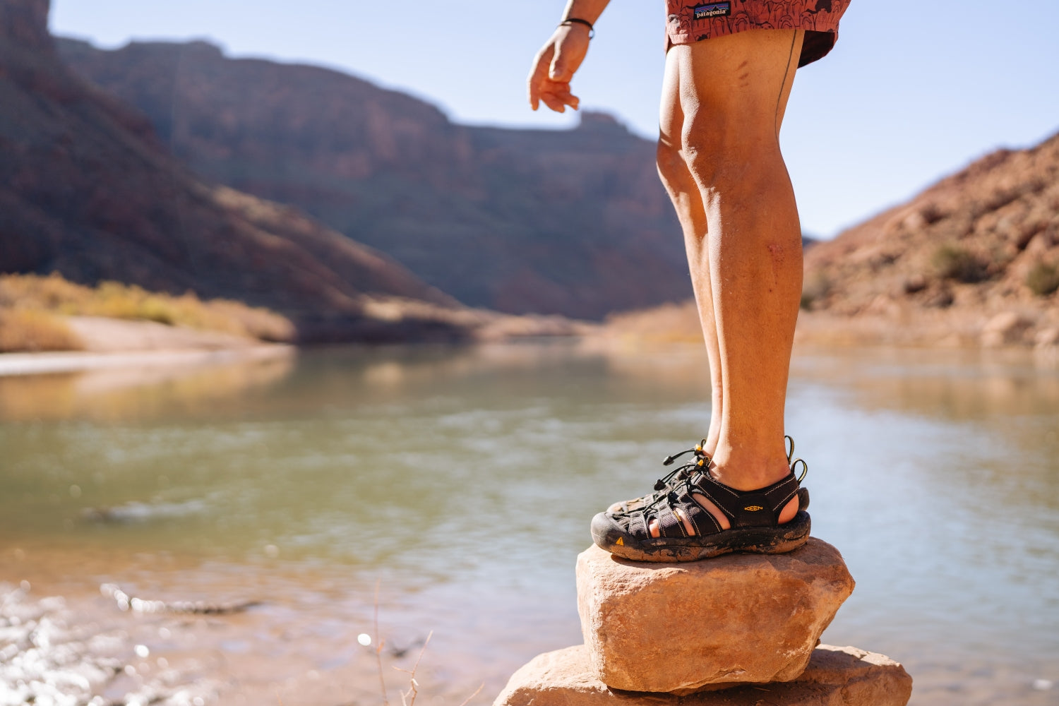 Dettaglio persona con sandali KEEN neri, in piedi su una roccia vicino a un fiume in canyon desertico.