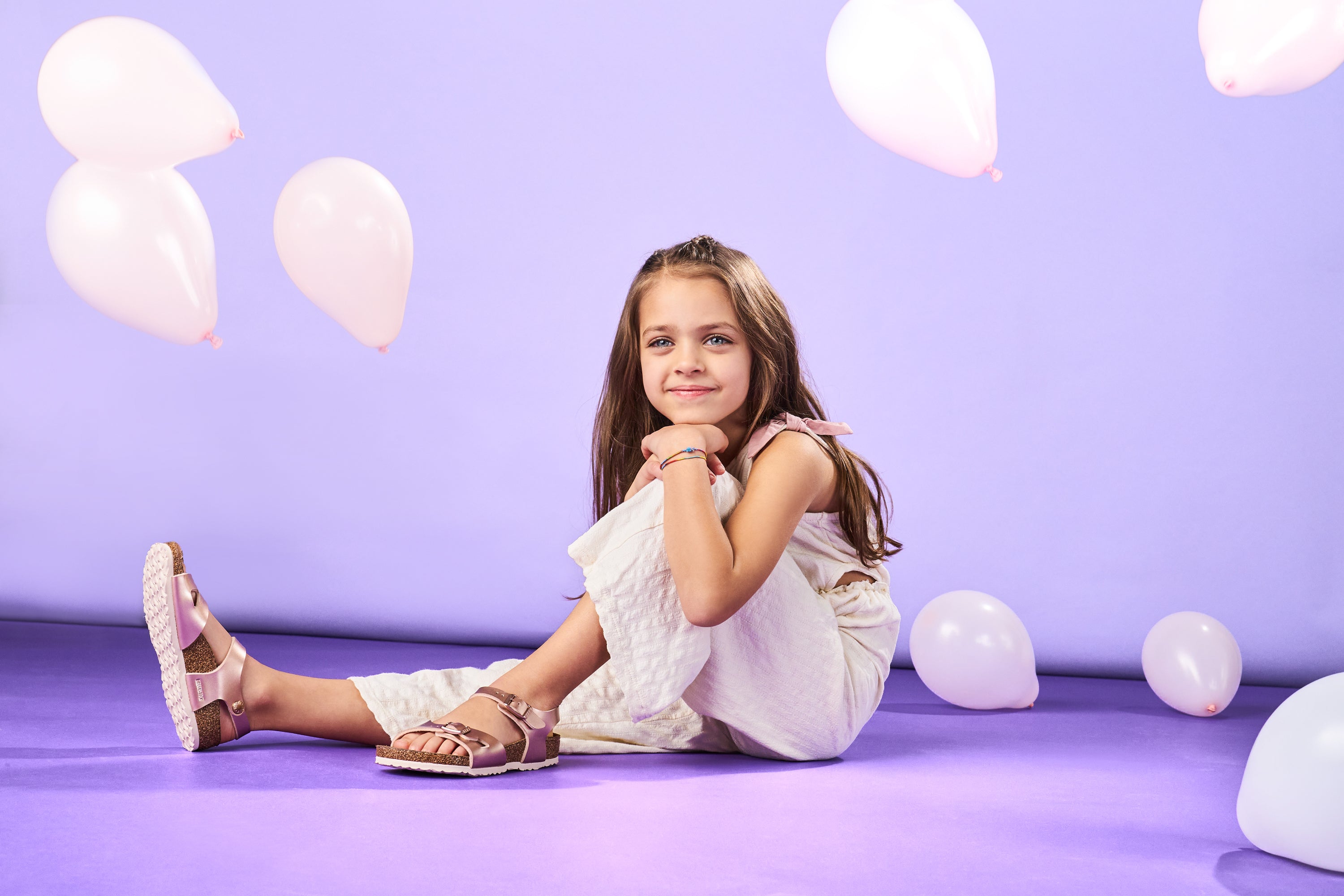 Bambina seduta su sfondo lilla con palloncini bianchi, indossa sandali Birkenstock Rio rosa.