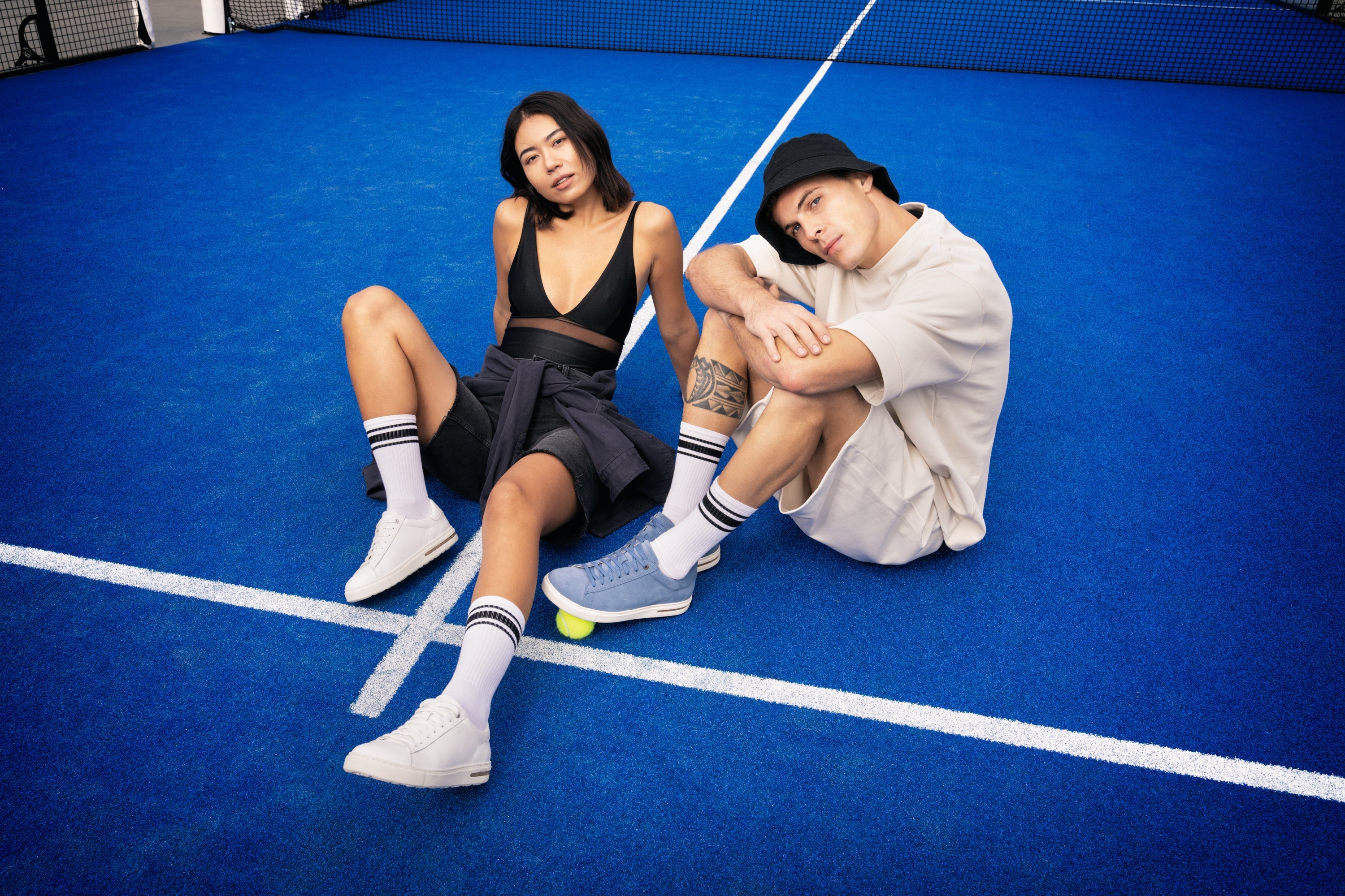 Due modelli seduti su un campo da tennis blu indossano sneakers Bend Low di Birkenstock: lei in pelle naturale bianca, lui in pelle scamosciata azzurra.