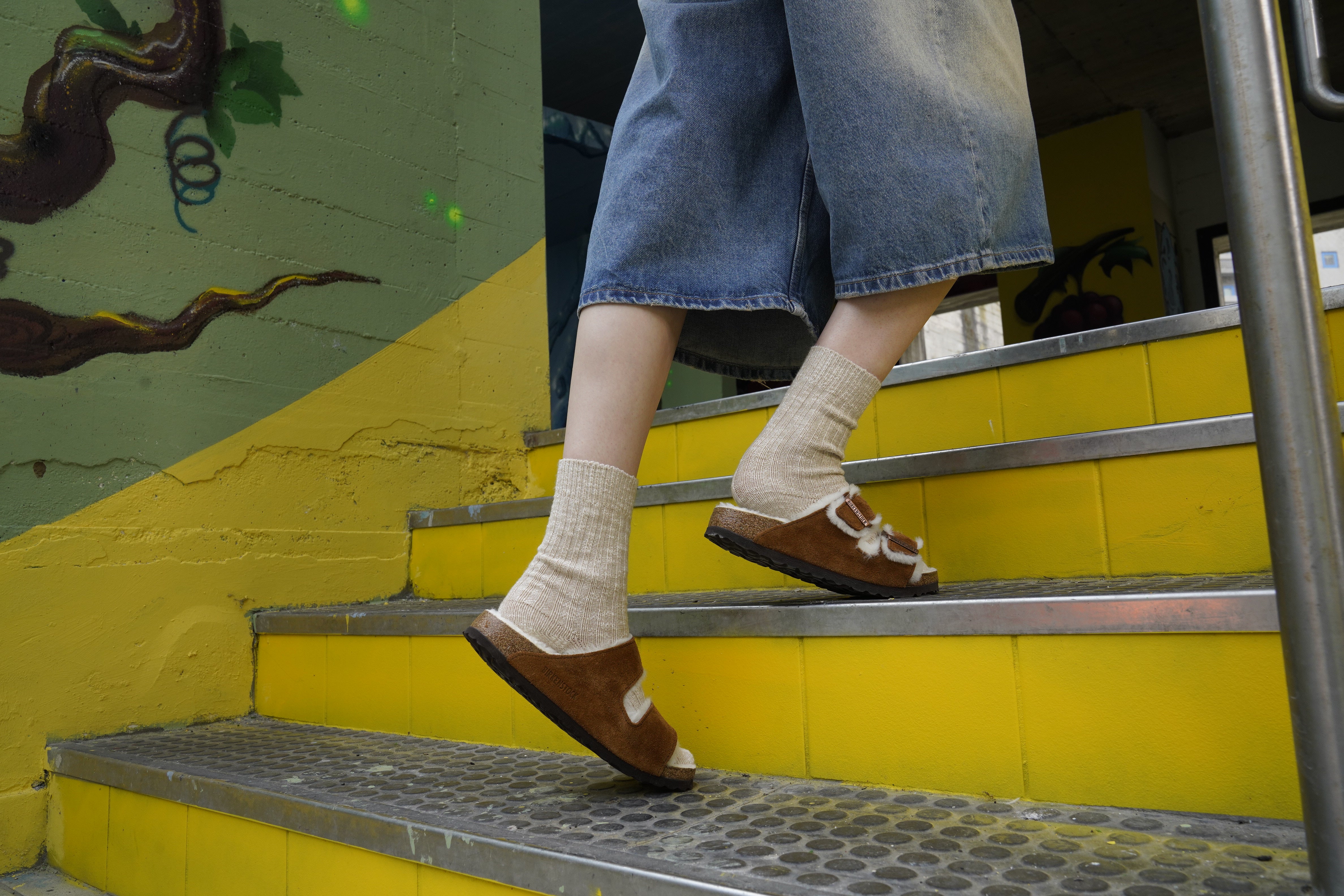 Persona che sale le scale indossando sandali Birkenstock Arizona Shearling Unisex color mink, in pelle scamosciata, abbinati a calzini beige e jeans cropped – ambientazione urbana con pareti colorate.