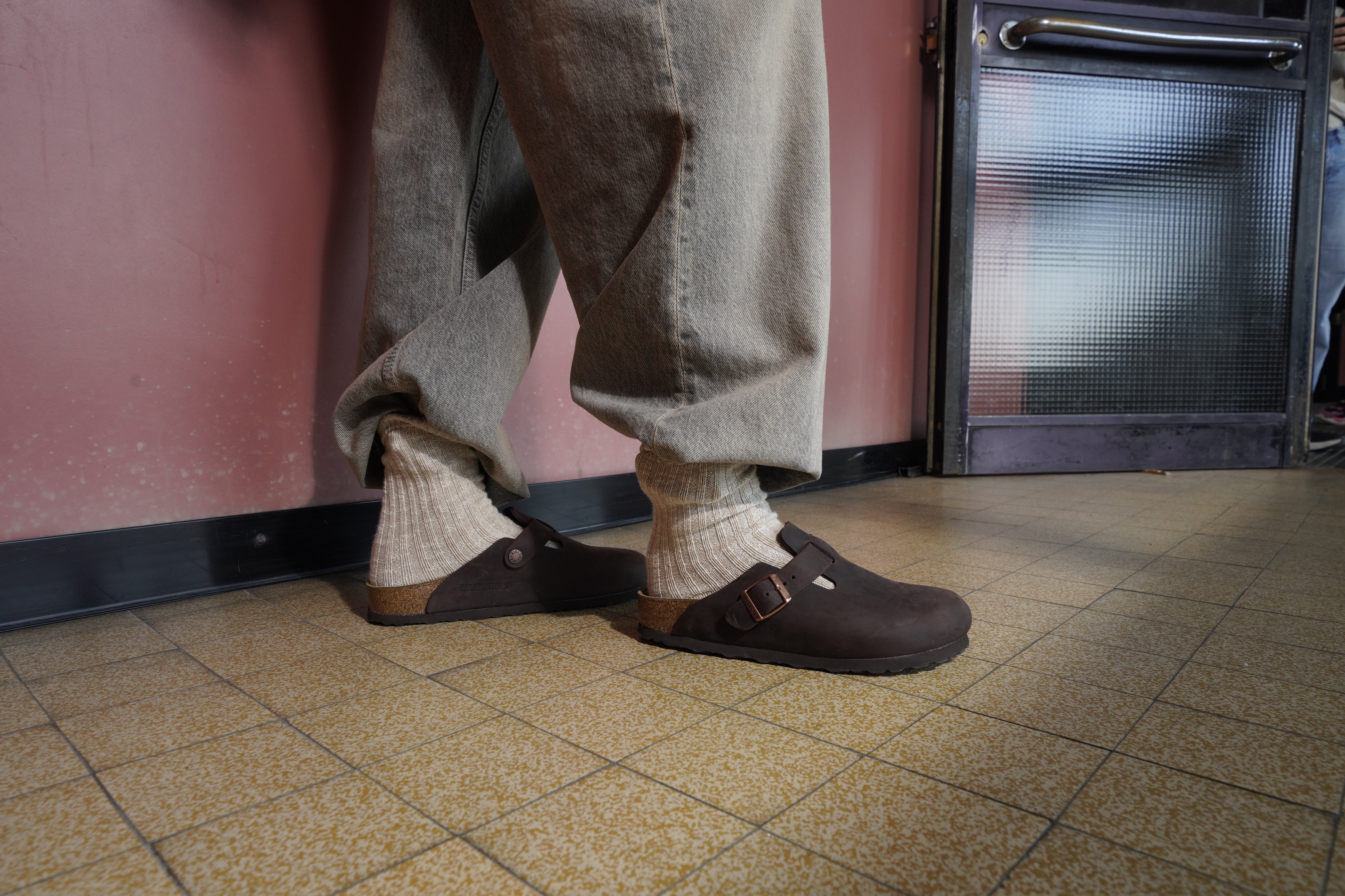 Primo piano di piedi che indossano Boston Sabot Unisex colore Habana in pelle ingrassata, con calzini chiari e pantaloni casual. Sullo sfondo, parete rosa e porta in vetro smerigliato, ambiente interno dallo stile retrò.