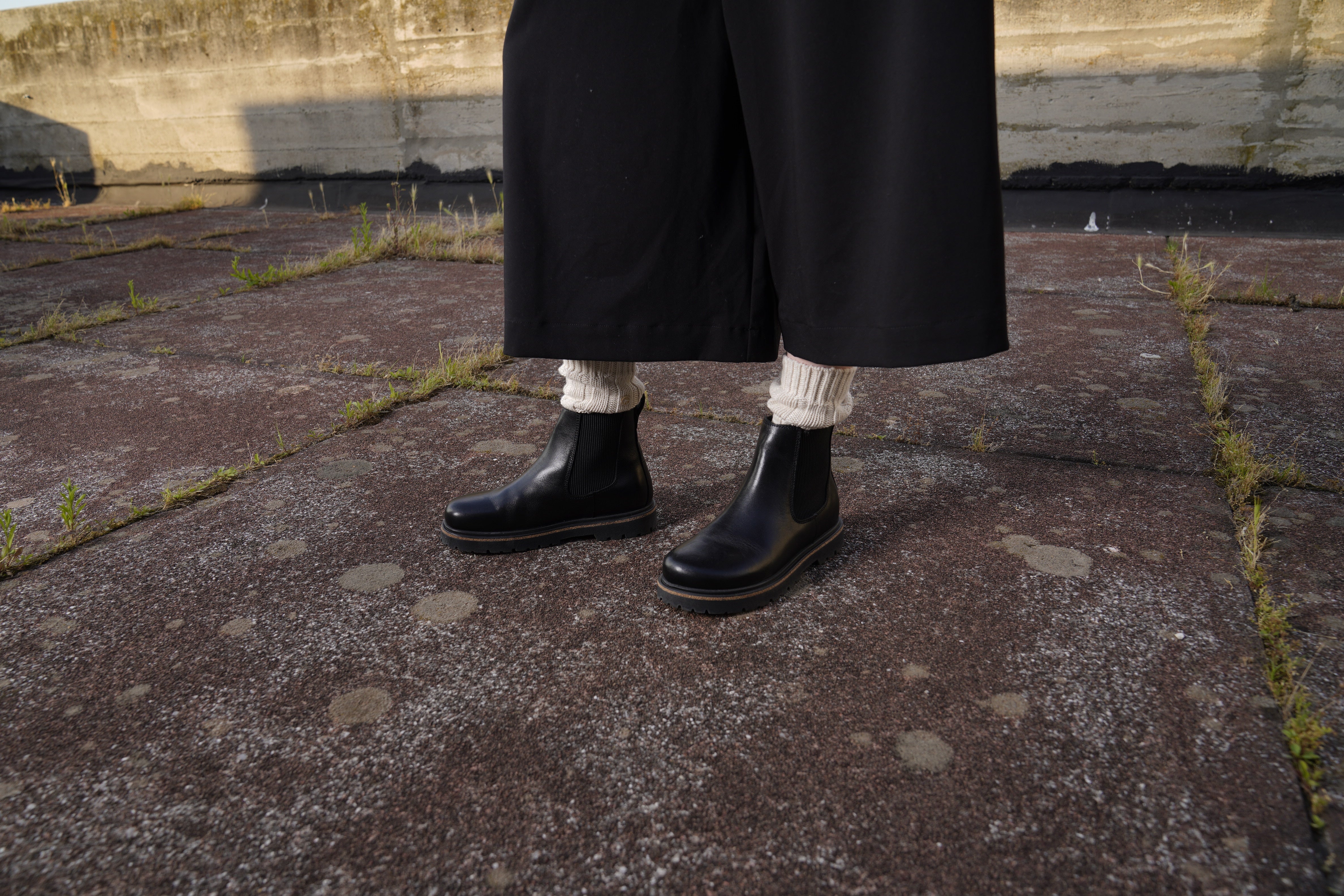 Persona con pantaloni neri ampi indossa gli stivaletti Birkenstock Highwood da donna in pelle bottalata nera, con design Chelsea, inserti elastici laterali e suola in TR antiscivolo. Calzatura ideale per l’autunno e l’inverno.