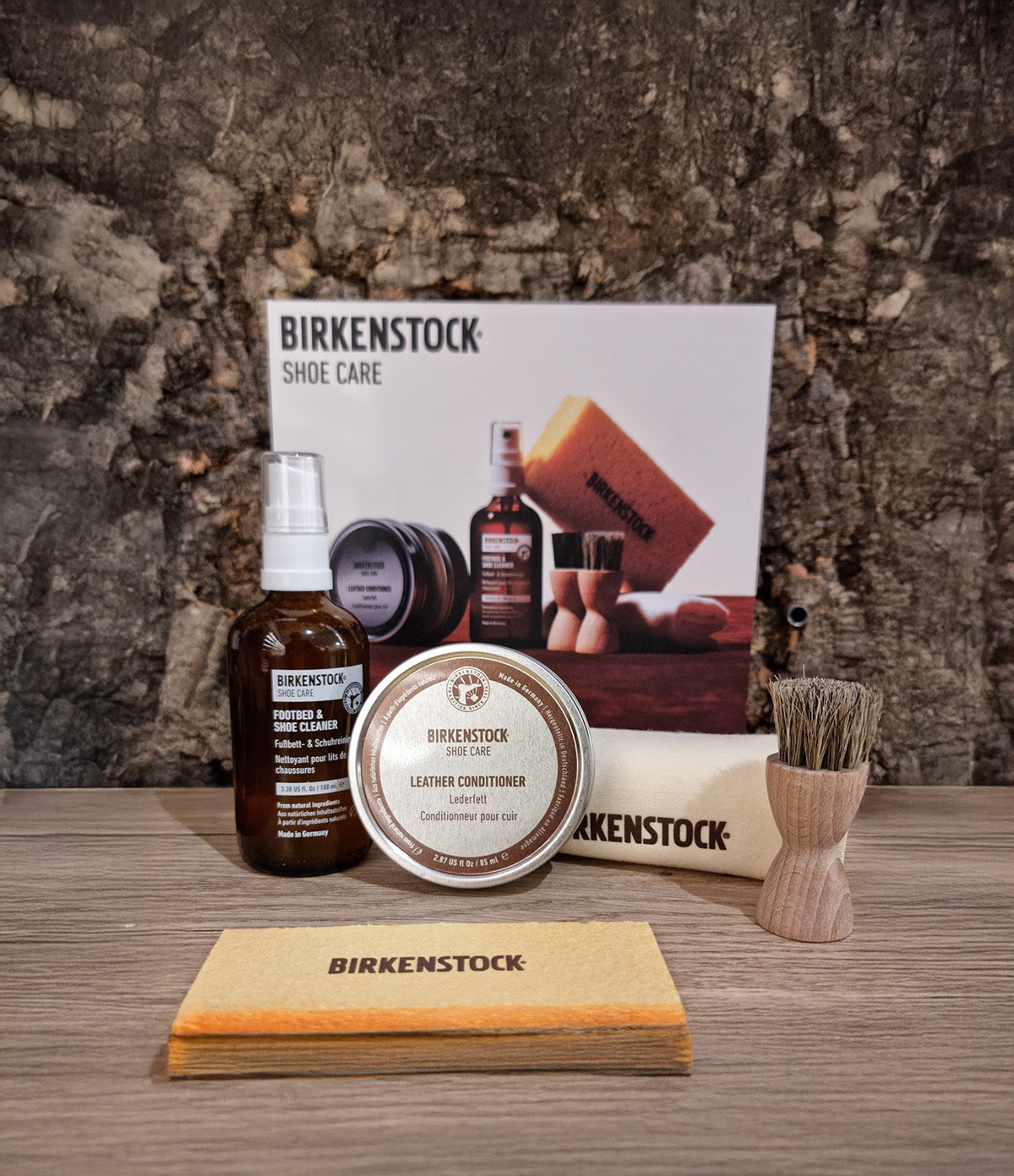 Set per la cura della pelle oliata Birkenstock composto da Leather Conditioner, detergente Footbed & Shoe Cleaner con spruzzatore, panno lucidante in cotone, spazzola in legno con setole naturali e spugna, esposti su tavolo in legno con fondo in pietra.