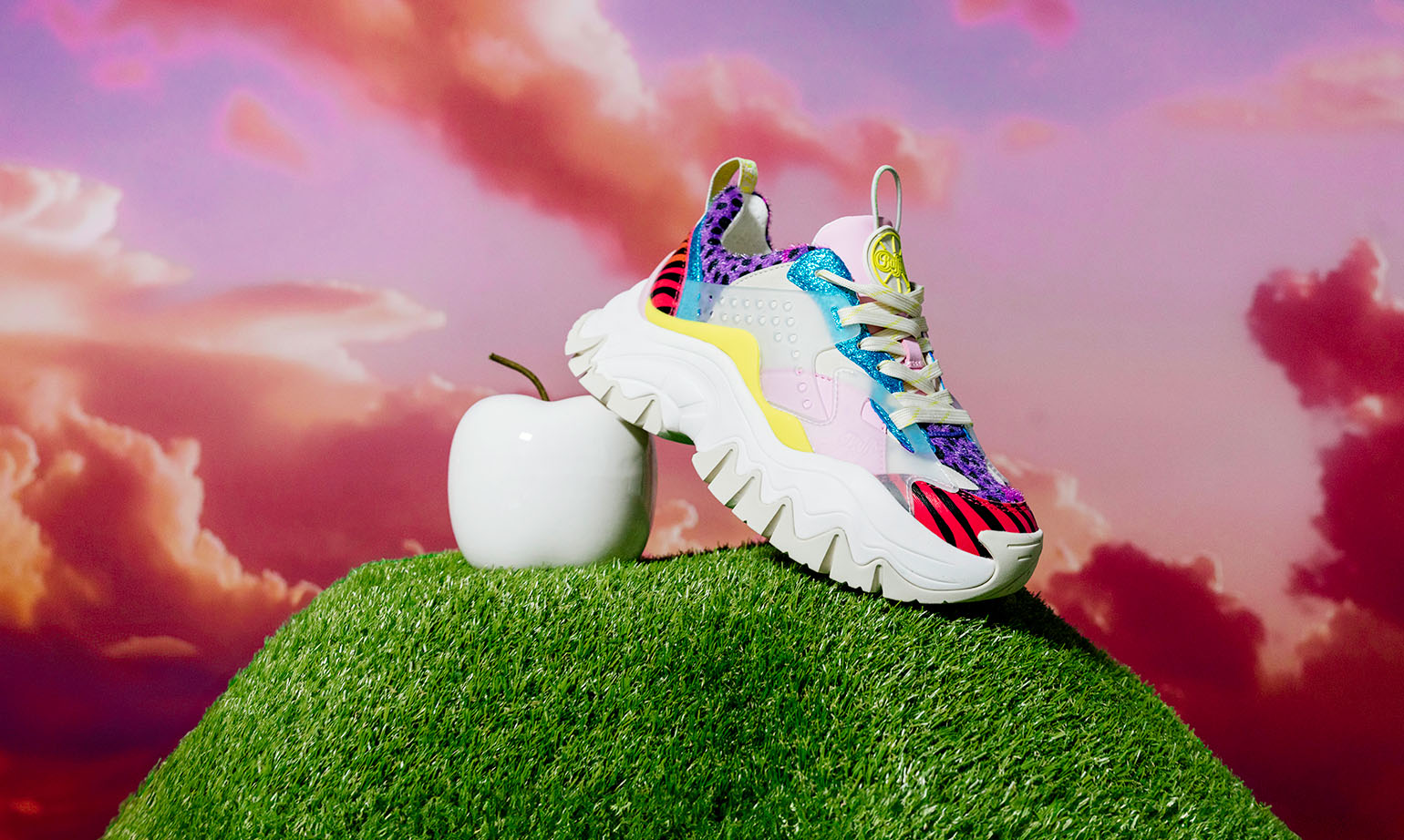 Scarpa Buffalo CLD Chai da donna, vegan e multicolore, con suola chunky, appoggiata su erba sintetica accanto a una mela bianca, con cielo rosa e nuvole sullo sfondo.
