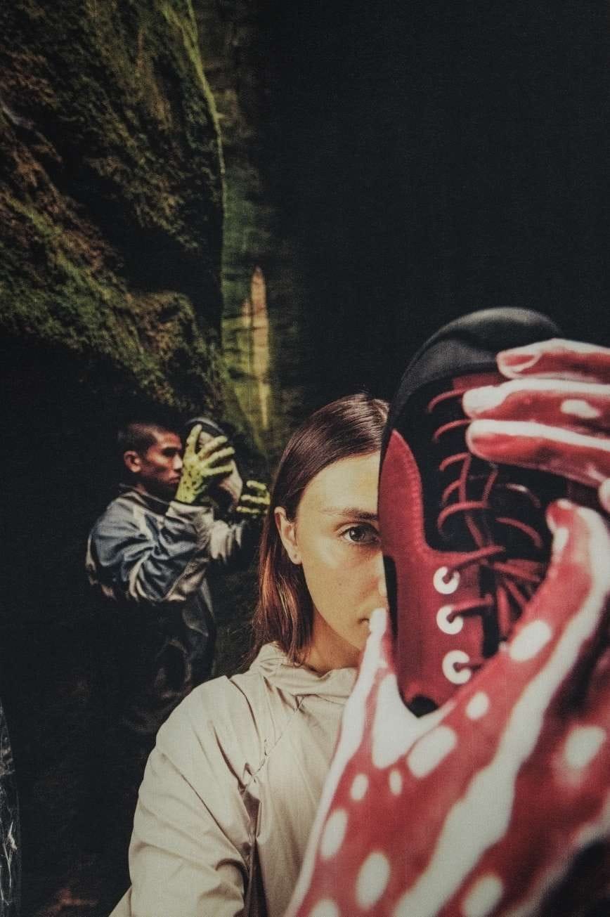 Una donna in primo piano tiene in mano una scarpa KEEN Jasper Rocks rosso scuro, mentre un uomo sullo sfondo mostra la versione color safari, entrambi all’interno di un canyon roccioso. Immagine della collaborazione tra KEEN e Hiking Patrol.