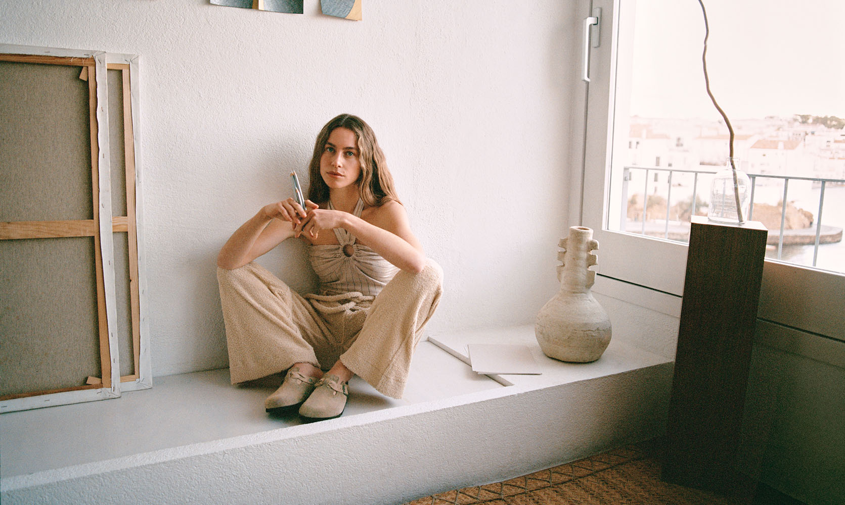 Donna seduta su un ampio davanzale interno accanto a una finestra, indossa sabot Birkenstock Boston Braided da donna in pelle scamosciata colore taupe.