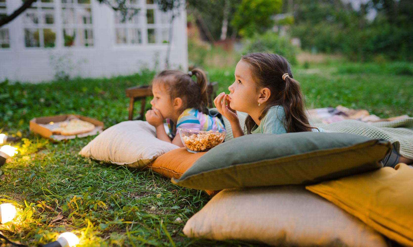 Due bambine sdraiate su cuscini in giardino mangiano popcorn, circondate da luci decorative.