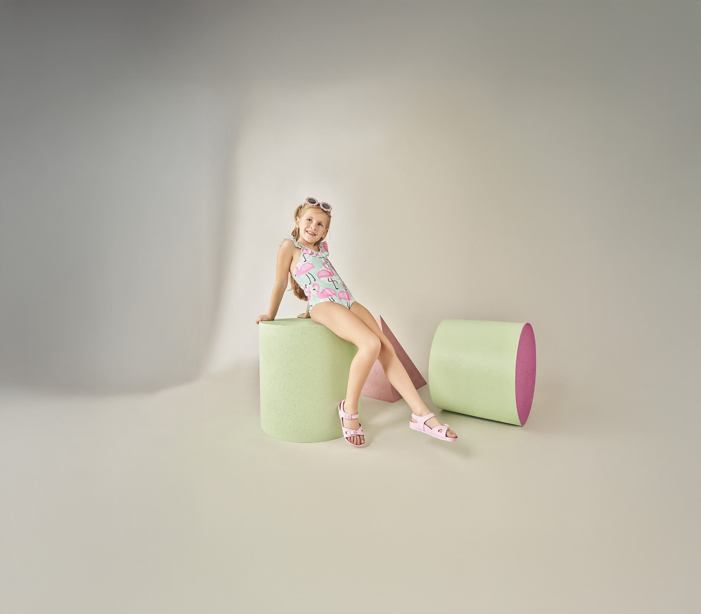 Bambina che indossa i sandali Birkenstock Rio EVA rosa, abbinati a un costume intero colorato, seduta su un cilindro verde pastello. Sandali leggeri, flessibili e impermeabili, ideali per l’estate.