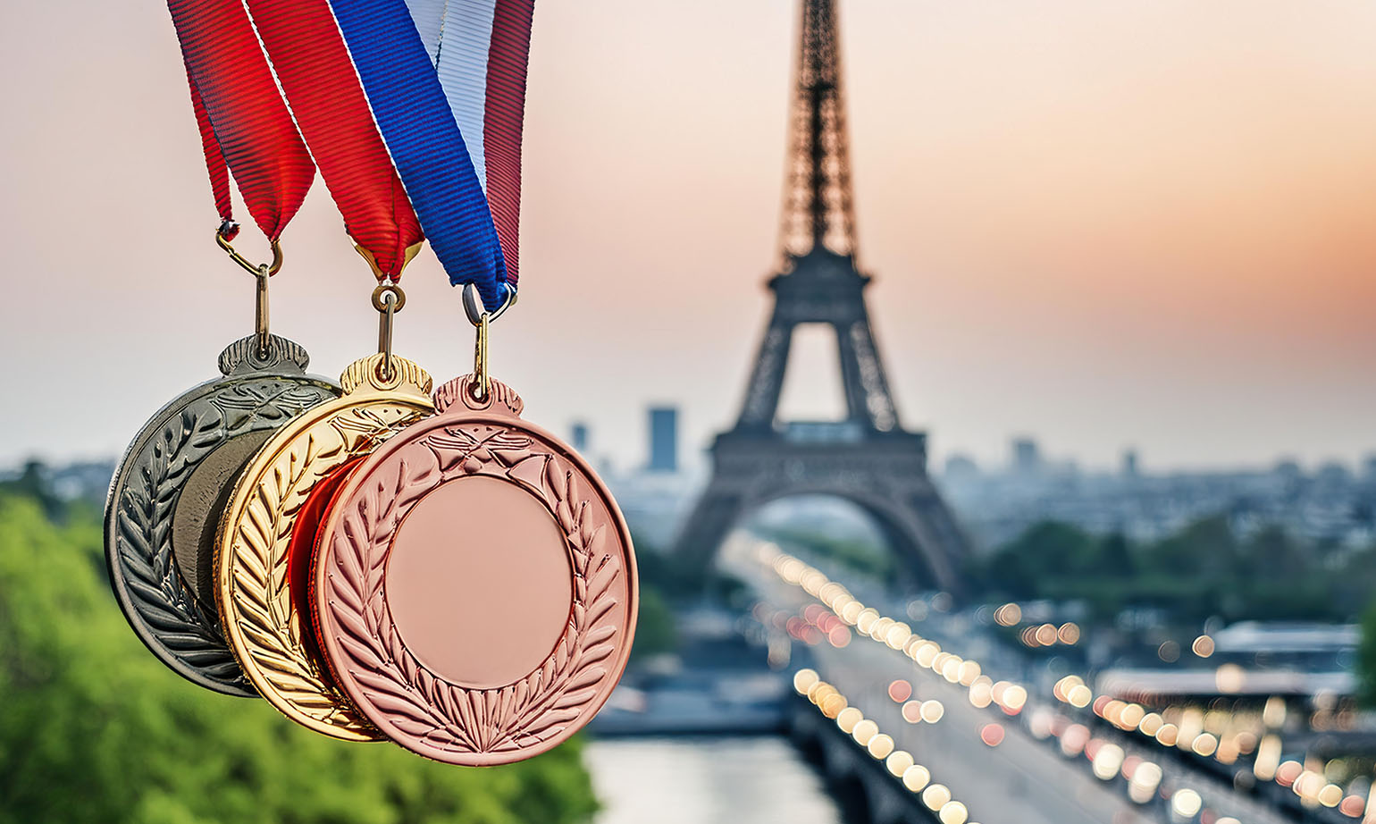 Medaglie olimpiche d’oro, argento e bronzo con nastri nei colori del tricolore francese, sospese in primo piano davanti alla Torre Eiffel al tramonto, in vista dei Giochi Olimpici di Parigi 2024.