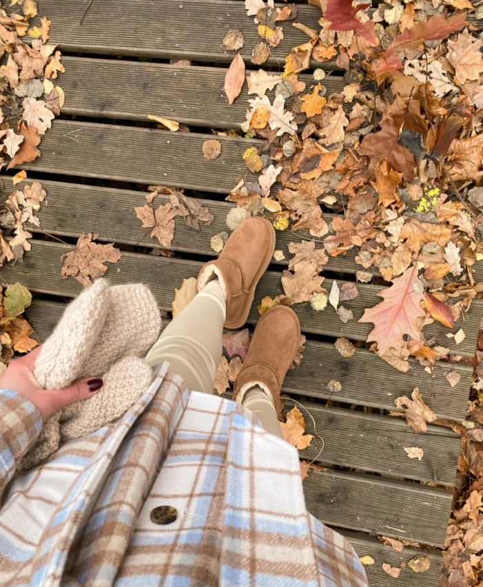 Vista dall’alto di una donna che indossa gli stivaletti EMU Australia Stinger Mini Donna color chestnut, in montone australiano double face resistente all’acqua, su passerella in legno coperta da foglie autunnali.