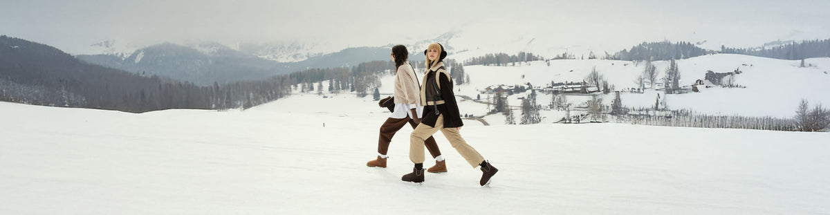 Donna seduta su sgabello in ambiente minimal con pareti chiare; indossa maglione crema, pantaloni bianchi e stivaletti EMU Australia Stinger Micro chocolate, riflessa in uno specchio