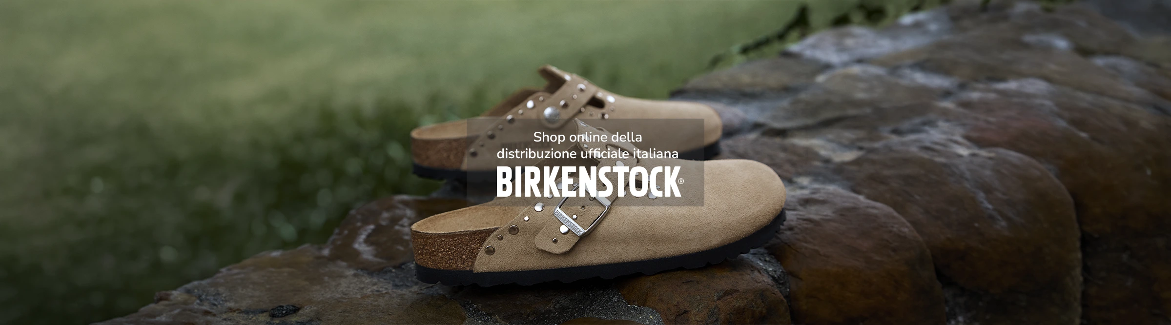 Un paio di sabot birkenstock modello boston con borchie in pelle scamosciata color taupe appoggiato su muro di pietra
