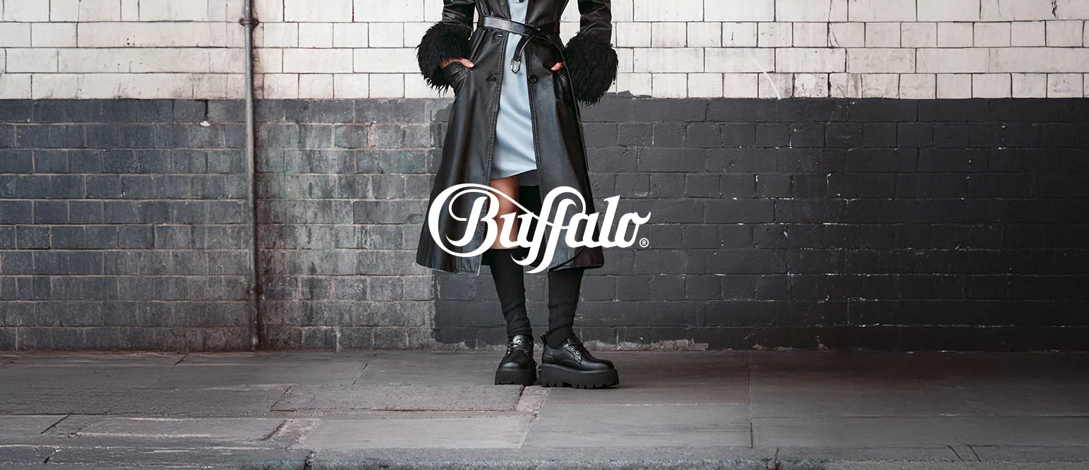 donna posa di fronte a un muro in contesto underground indossando scarpe buffalo nere con calze nere fino al ginocchio, vestito azzurro e cappotto lungo di pelle nera