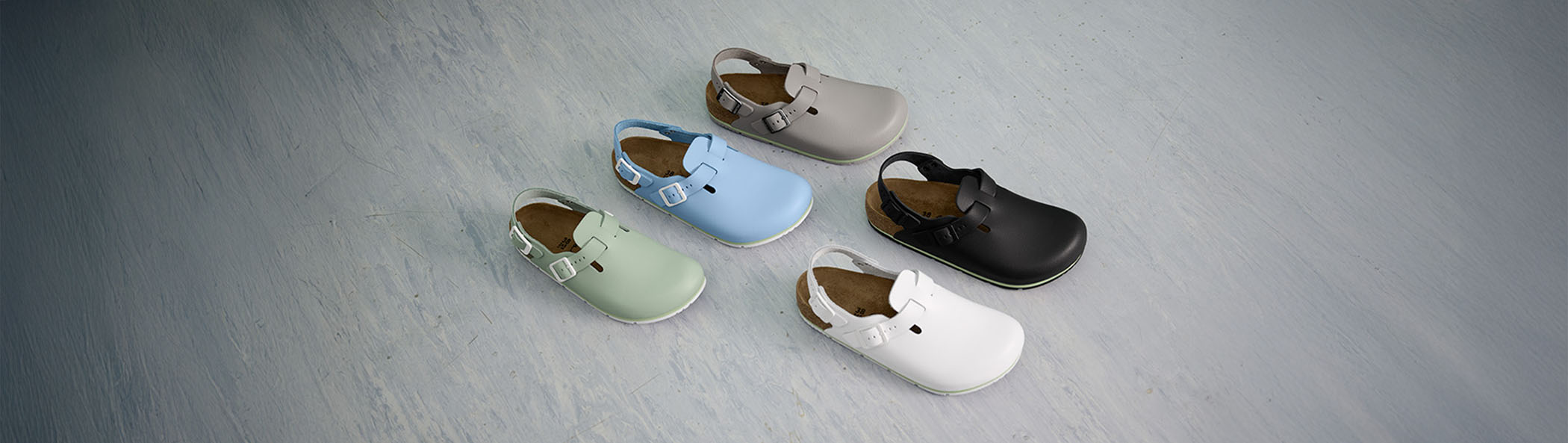 Cinque paia sabot Birkenstock Professional, ideali per uso professionale in settori come HORECA, sanitario e industriale, disposti su una superficie grigia che ricorda un pavimento. I sabot sono visti dall'alto e leggermente di lato, mettendo in risalto le diverse colorazioni e il design classico del modello.