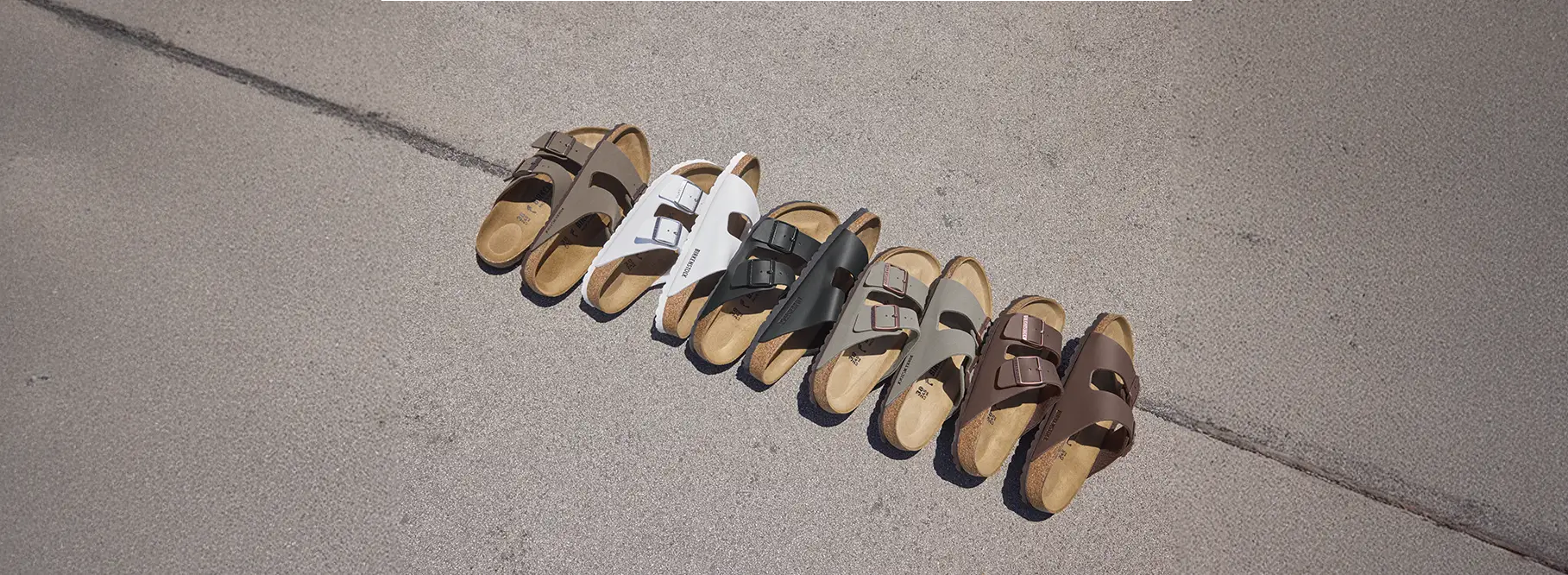 Cinque paia di ciabatte sandali Arizona di Birkenstock di varie colorazioni (mocca, bianche, grigie, marroni scuro e nere) appoggiate su pavimento in vista dall'alto