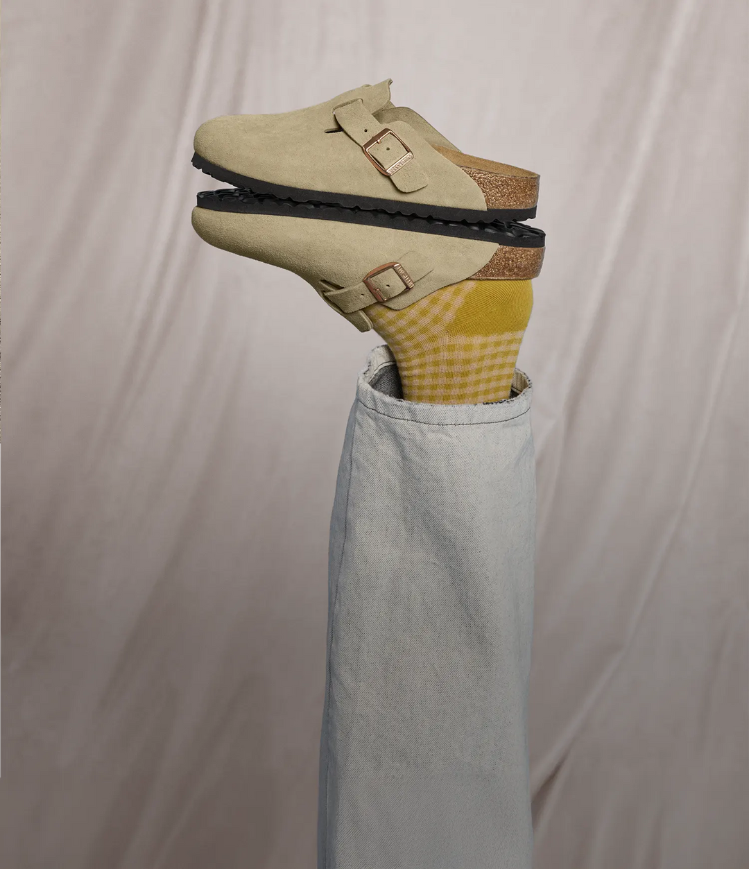 Vista laterale di due birkenstock sabot birkenstock boston taupe, impilati l'uno sull'altro. Un piede con calzini a quadri gialli e bianchi è posizionato all'interno del sabot rivolto verso il basso