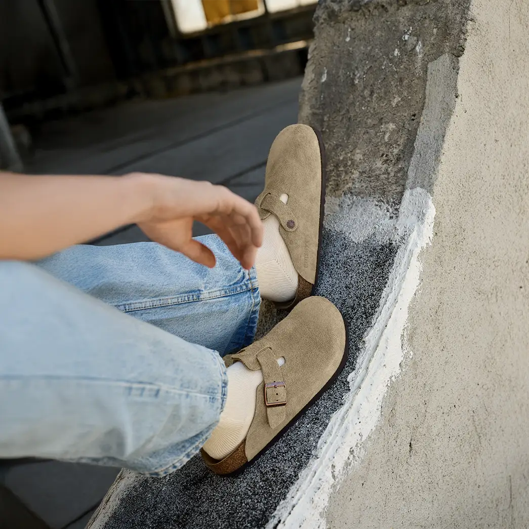 primo piano di persona con indosso boston sabot birkenstock taupe con indosso calzini bianchi e jeans