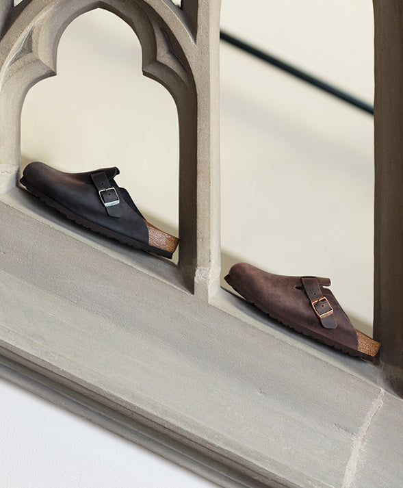 Primo piano laterale di due paia di Birkenstock boston clogs in pelle oliata, una nera e una habana marrone appoggiate sul cornicione di una scala in pietra