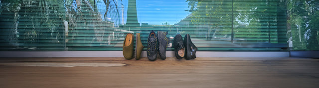 Diverse calzature Amuvia — pantofole Haflinger, scarpe KEEN e FinnComfort — disposte su un pavimento in legno davanti a una vetrata, simbolo di comfort, qualità e design per ogni stagione.